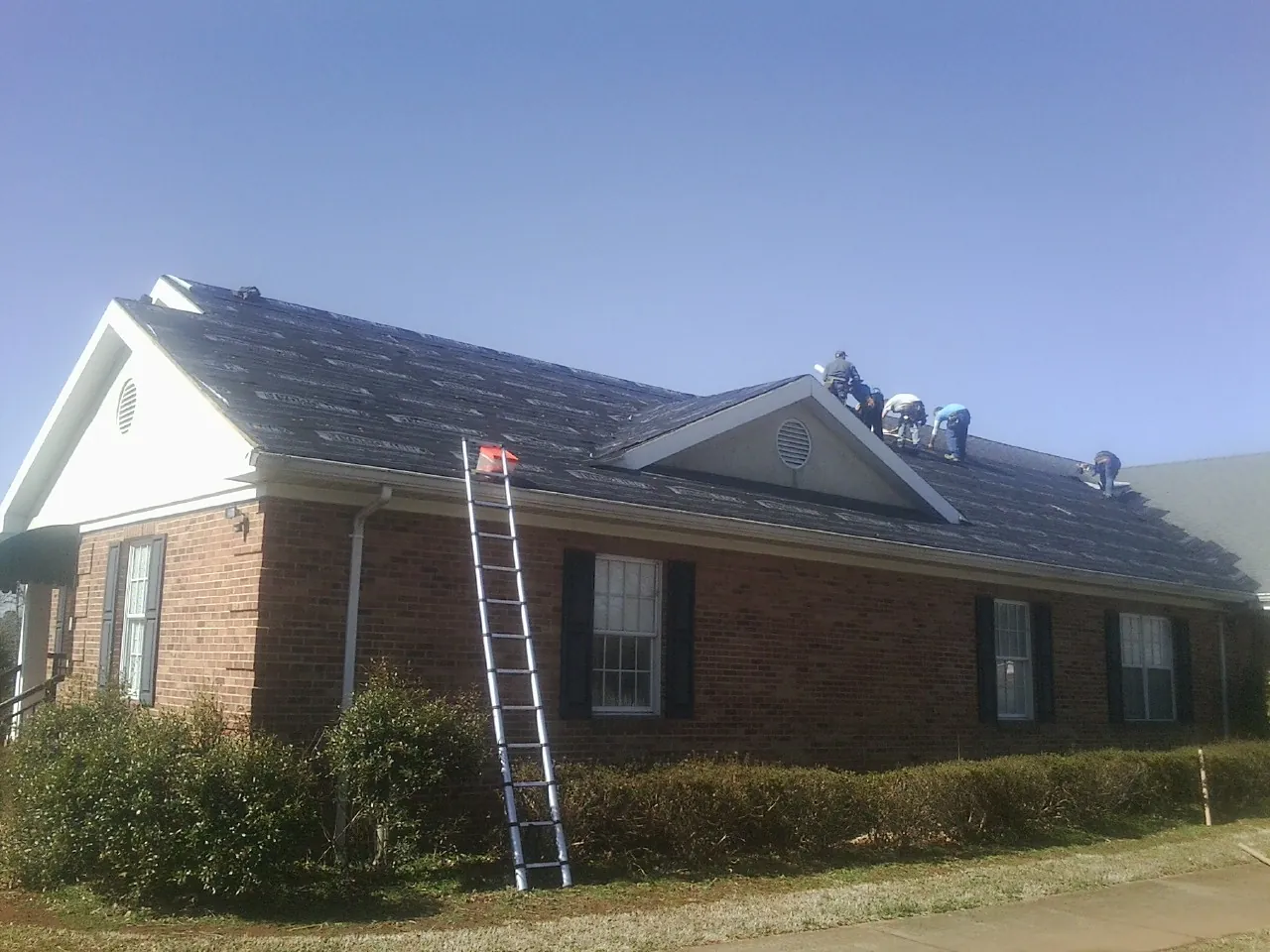 Crew tearing shingles off a brick ranch roof with a ladder set — Emergency Roof Repair work by our crew in Eden
